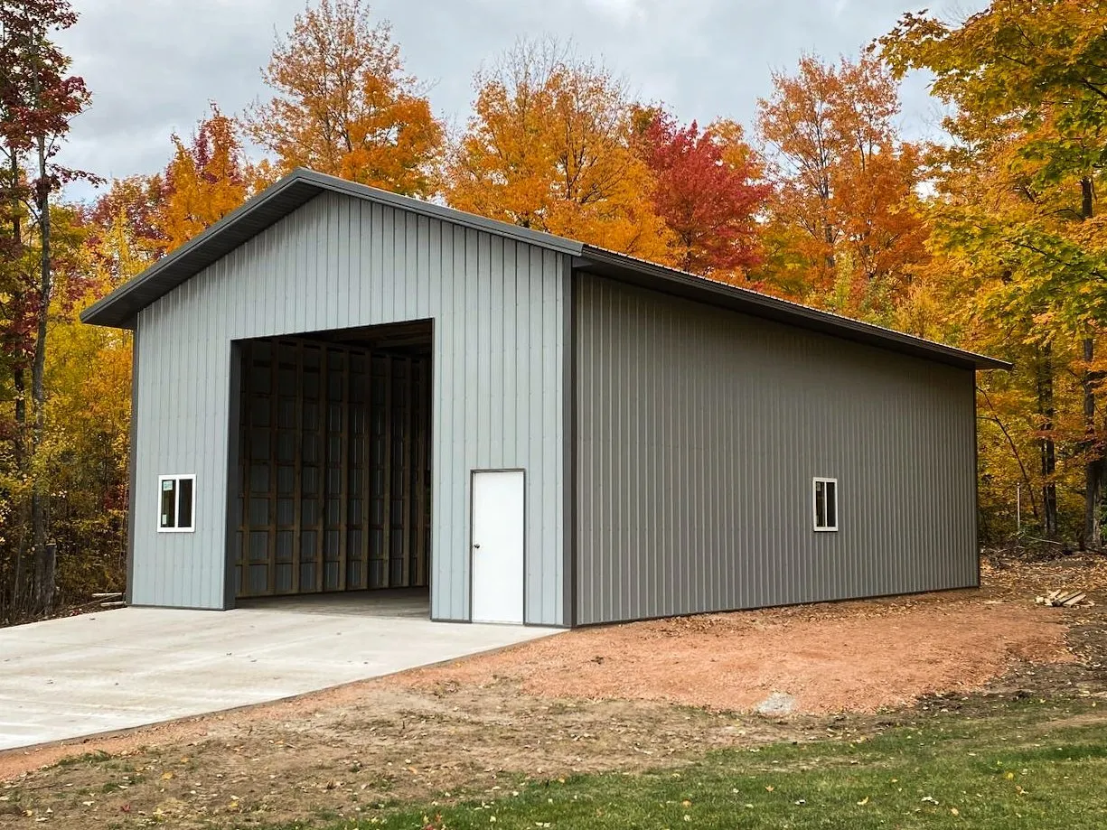 Grey Metal Garage with Open Bay A complete grey metal garage with white trim, featuring one large open bay and a side entry door, set in a yard with bright autumn foliage in the background.