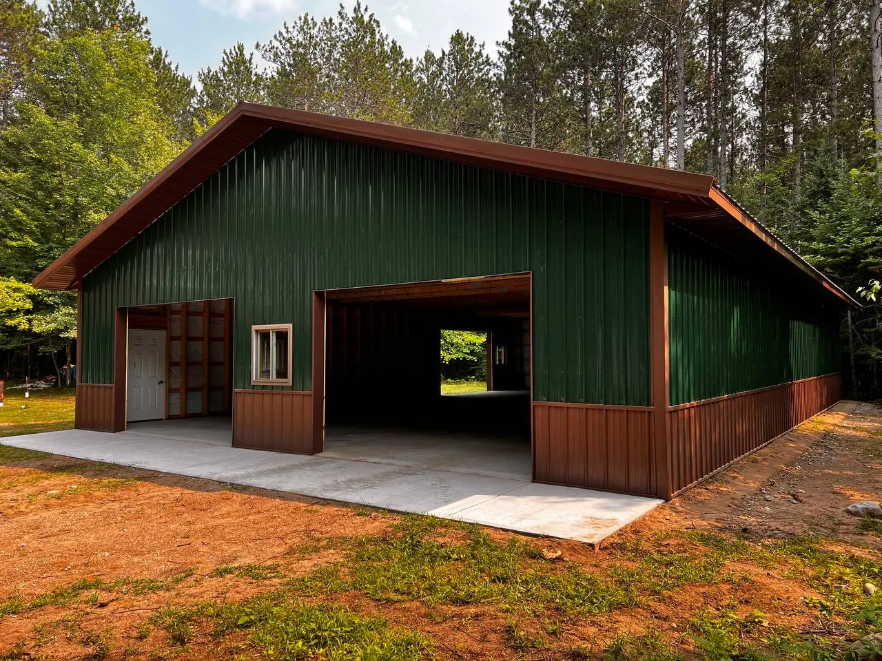 Large Green Metal Workshop with Brown Wainscot A wide exterior view of a durable green and brown metal workshop building with two open garage doors, constructed on a concrete foundation in a wooded clearing.