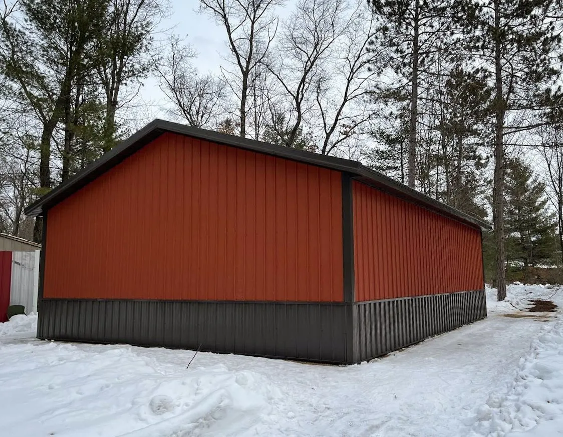 Red and Grey Metal Pole Barn in Snowy Landscape Exterior view of a finished red metal building with a charcoal grey wainscot panel, positioned against a snowy field and bare winter trees.